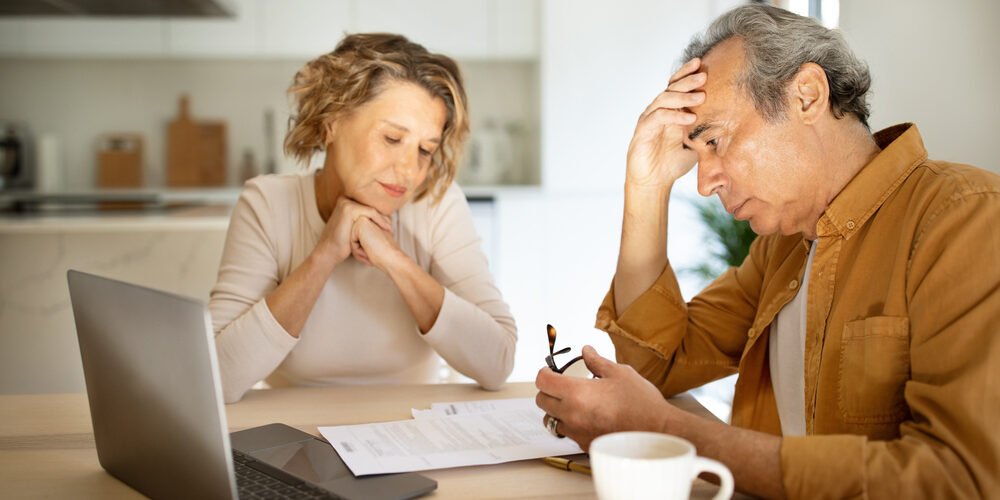 Stressed elderly couple checking financial papers, sitting with laptop and looking at loan documents, kitchen interior Financial advisor explaining retirement account division to a couple during a divorce.