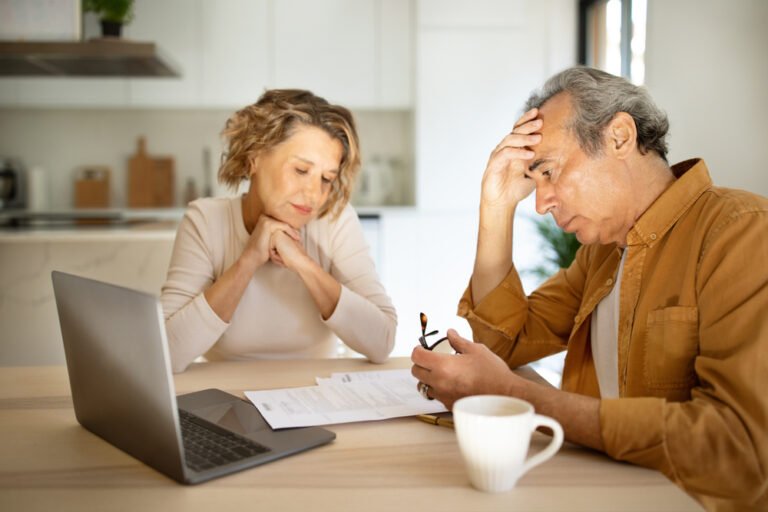 Stressed elderly couple checking financial papers, sitting with laptop and looking at loan documents, kitchen interior Financial advisor explaining retirement account division to a couple during a divorce.